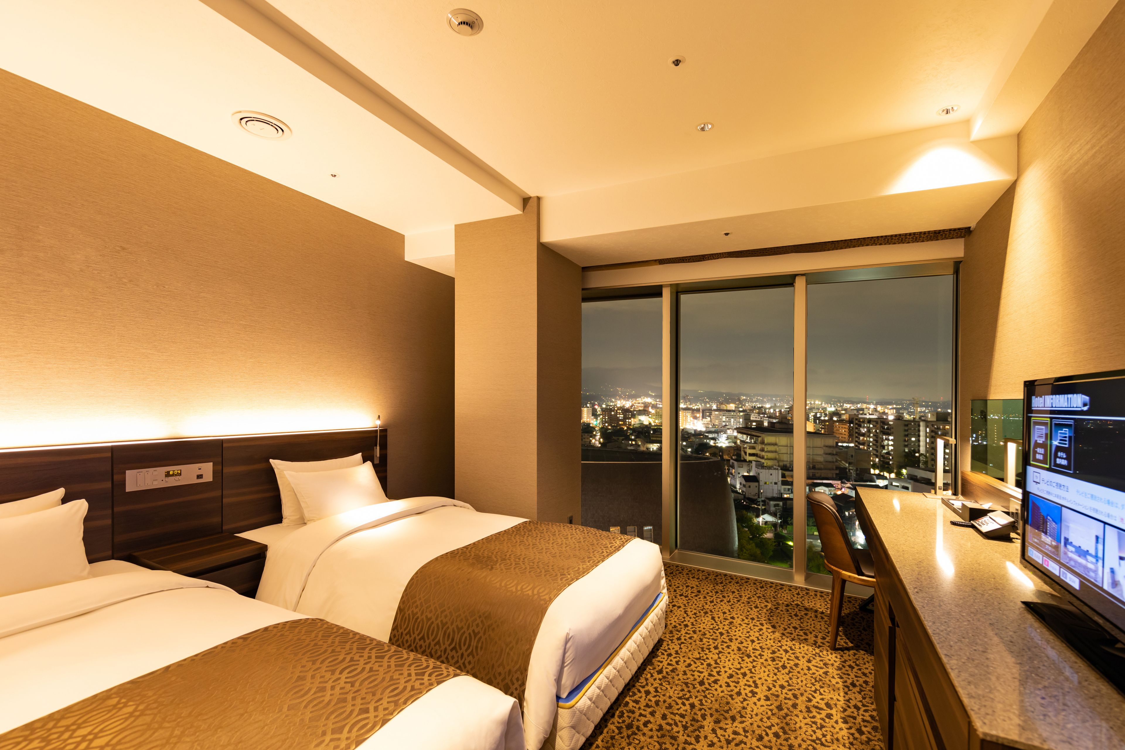 Habitación de hotel con dos camas, un escritorio, un televisor y un amplio ventanal con vista a la ciudad.
