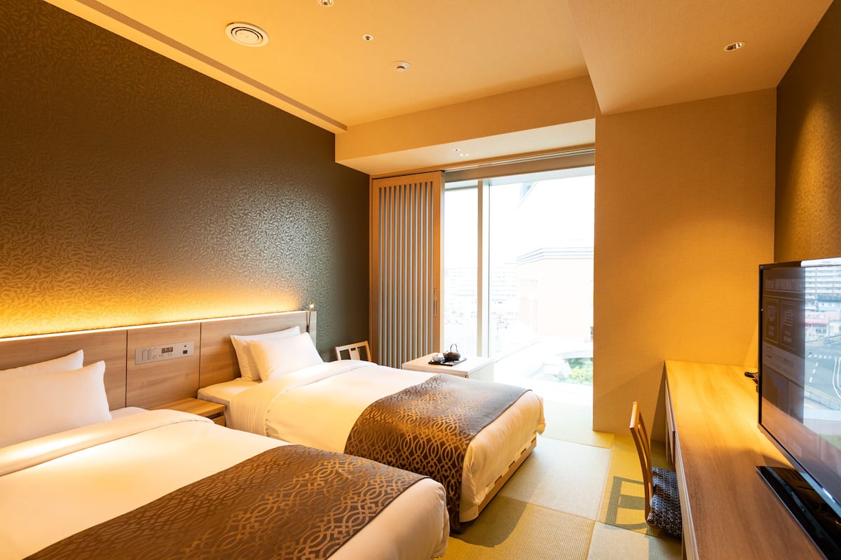 A hotel room with two beds, a TV, a small table, and a window with curtains.