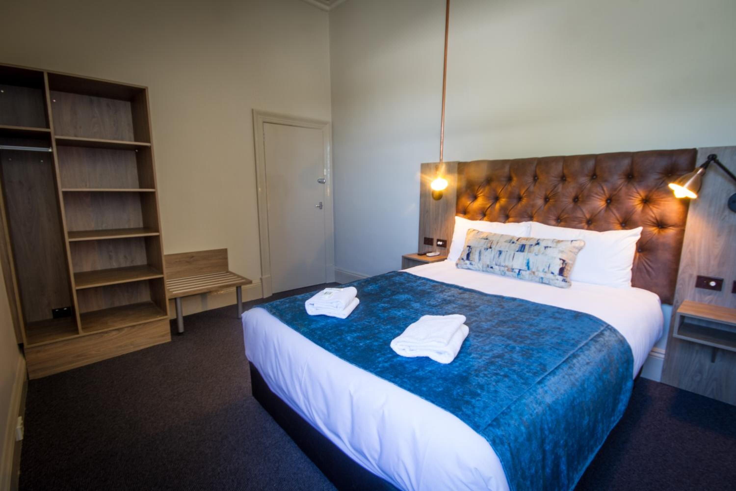 A hotel room with a large bed, a wooden bench, a wardrobe, and a bedside table with a lamp.