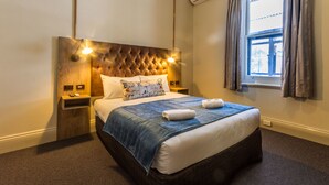 A hotel room with a tufted headboard, a large bed with a blue blanket, two pillows, a bedside table with a lamp, and a window with curtains. - Pretoria Hotel (Mannum)