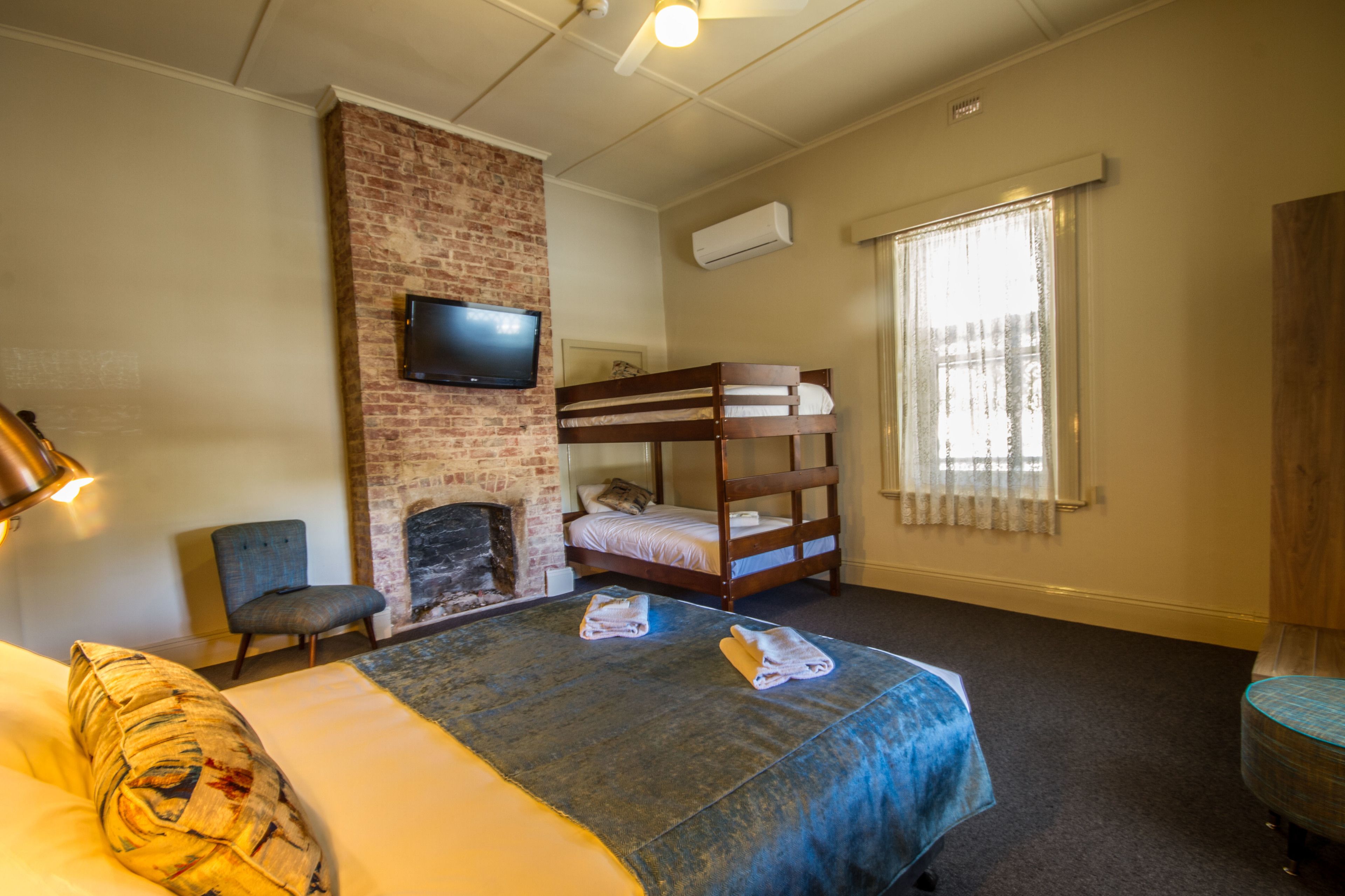 A room with a bed, a chair, a television mounted on a brick wall, a window with curtains, and a fireplace.