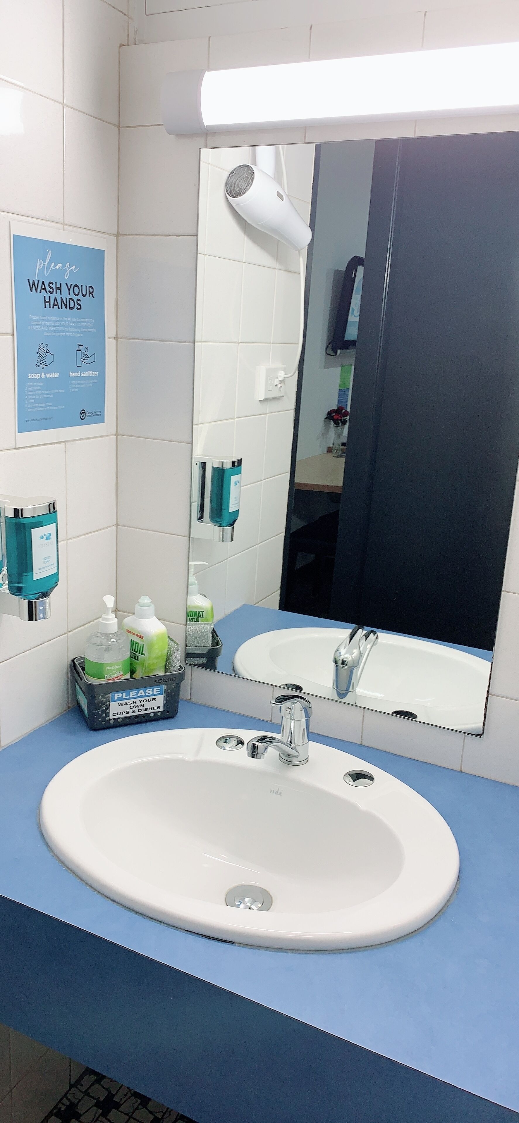 A bathroom with a white sink, blue countertop, and a mirror. There is a sign instructing to wash hands, a hairdryer, and hand sanitizer on the wall.