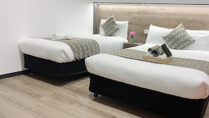 A hotel room with two beds, each with a wooden headboard, white bedding, and patterned throw pillows. There are small tables with decorative vases and a wooden nightstand. - Elm Motel (Golden Square)
