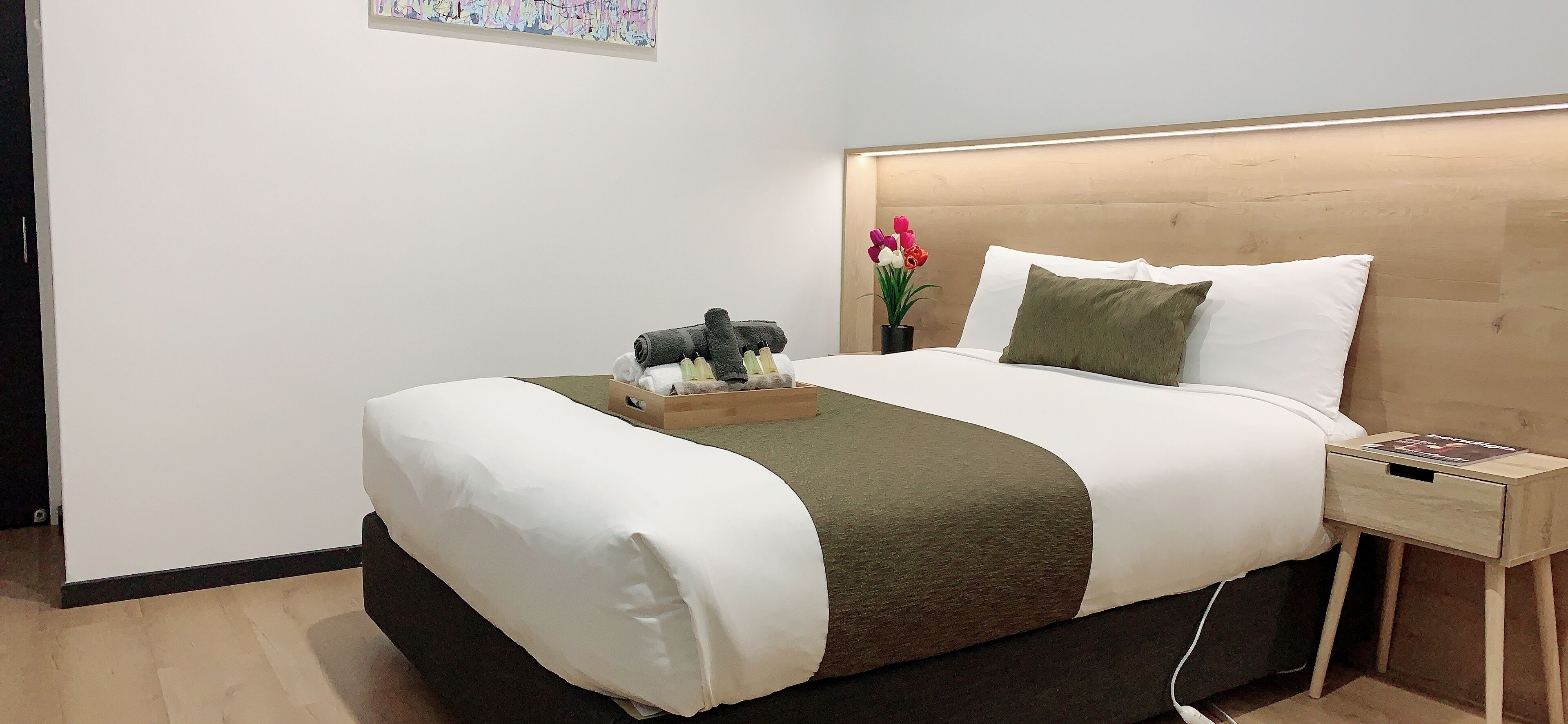 A neatly made bed with a wooden headboard, a bedside table with a magazine, and a small potted plant with flowers.