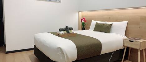 A neatly made bed with a wooden headboard, a bedside table with a magazine, and a small potted plant with flowers.