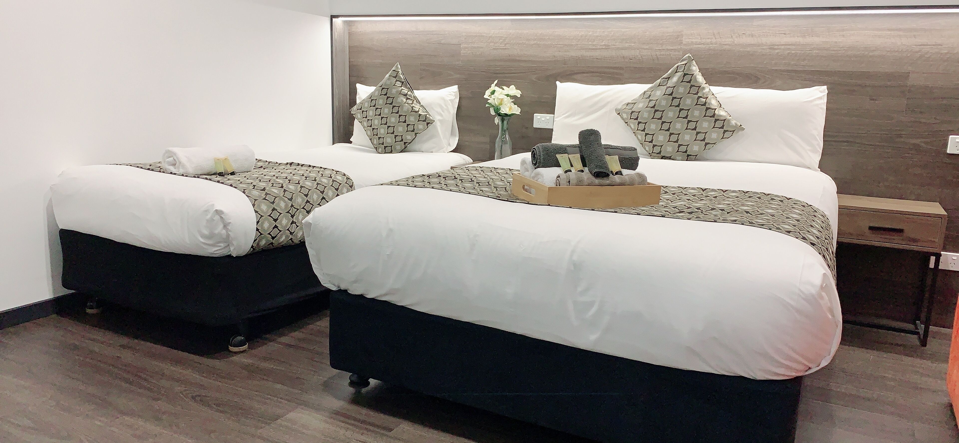 A hotel room with two beds, each with a pillow and a rolled towel. There is a wooden headboard, a nightstand, and a vase with flowers.