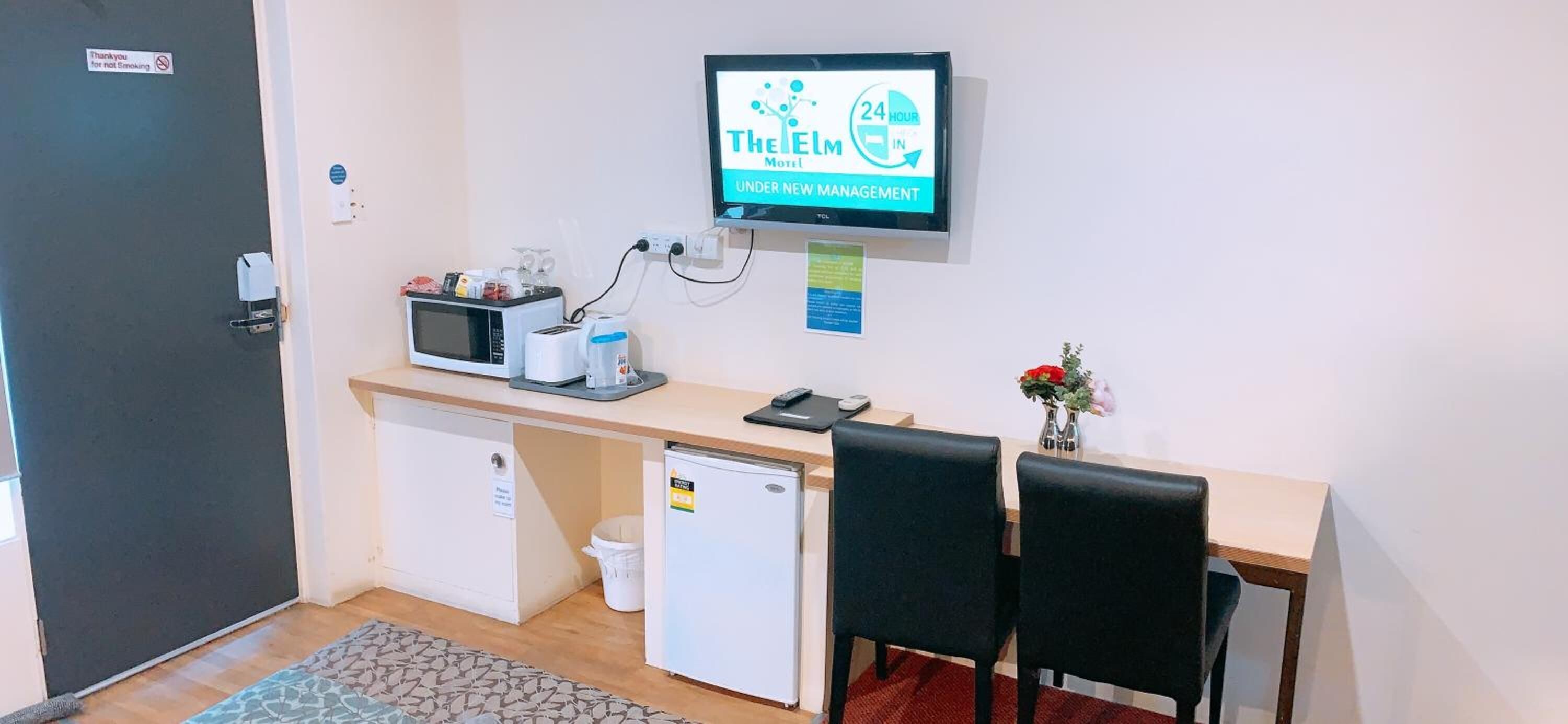 A small kitchenette with a microwave, toaster, and coffee maker, a TV displaying 'The Elm Motel' sign, a small fridge, and a desk with two chairs.