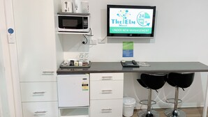 A kitchenette with a microwave, coffee maker, and a digital screen displaying hotel information. There are two bar stools and a trash bin. - Elm Motel (Golden Square)