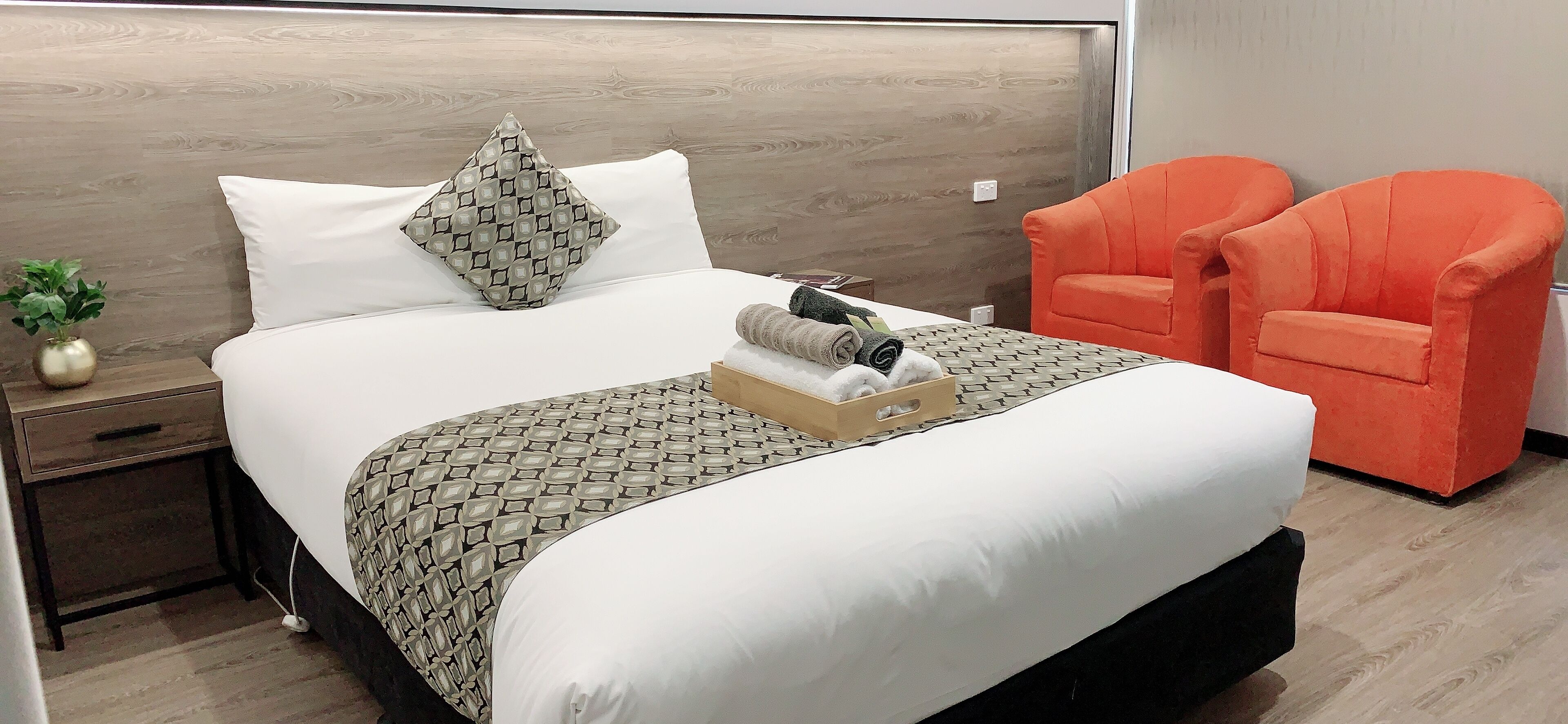 Una cama bien tendida con una colcha estampada, una almohada y una bandeja con toallas. Hay dos sillas color naranja brillante junto a la cama.