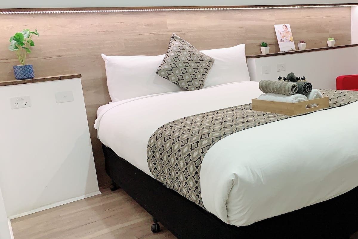 Ein ordentlich bezogenes Bett mit einem gemusterten Überwurf, ein zusammengerolltes Handtuch und eine Ablage mit Pflegeprodukten. Darüber befindet sich eine Holzablage mit zwei roten Stühlen und einer kleinen Topfpflanze.