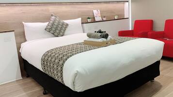 Una cama bien hecha con una colcha estampada, una toalla enrollada y una bandeja con artículos de tocador. Hay dos sillas rojas y una pequeña planta en maceta sobre una repisa de madera.