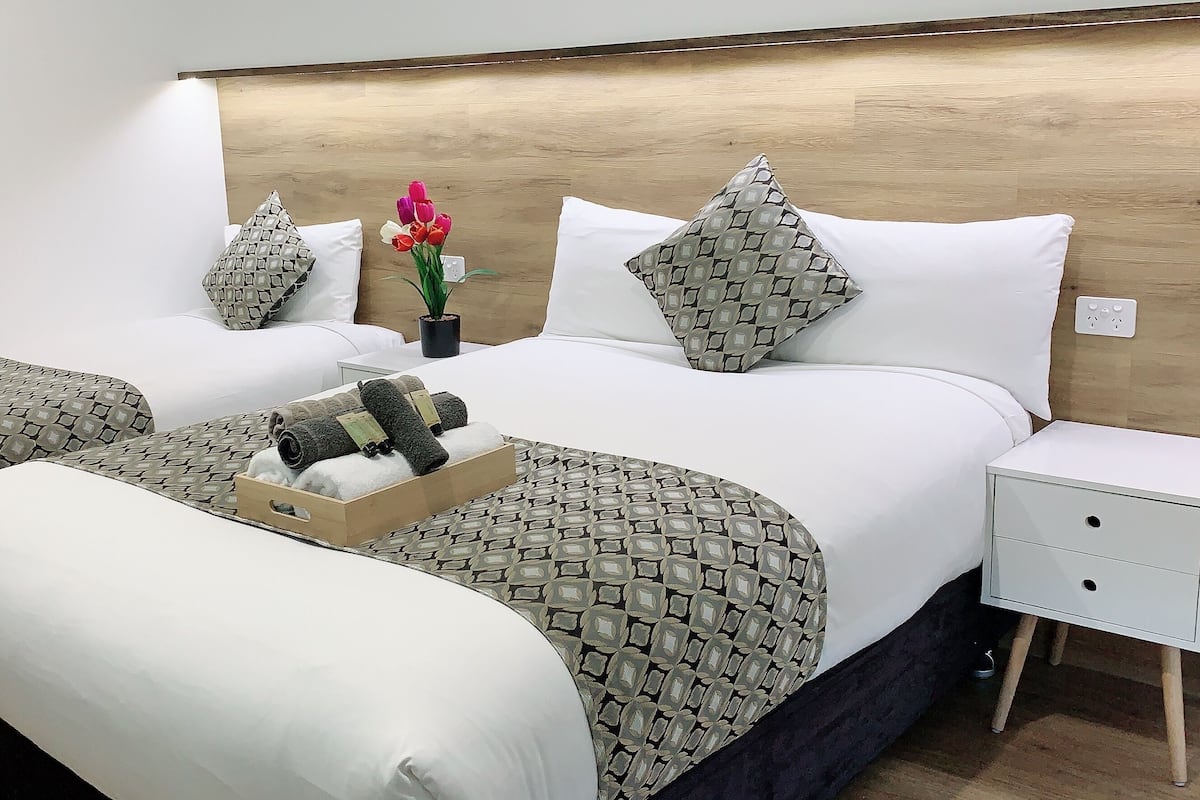 Ein Hotelzimmer mit zwei Betten, einem Holzkopfende, einem Nachttisch mit Schubladen und einem Tablett mit Handtüchern und Toilettenartikeln.