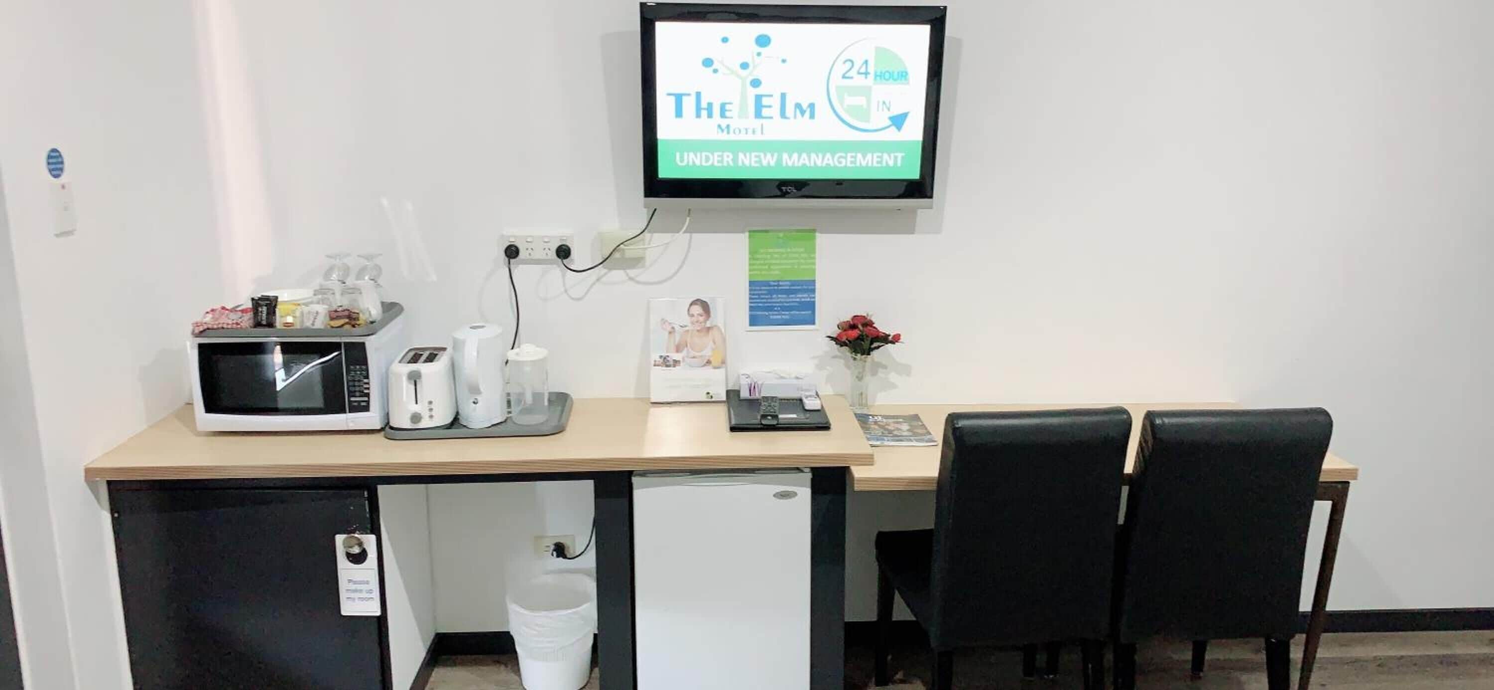 A hotel room with a desk, microwave, coffee maker, and a TV displaying 'The Elm Motel 24 Hour In Under New Management'.