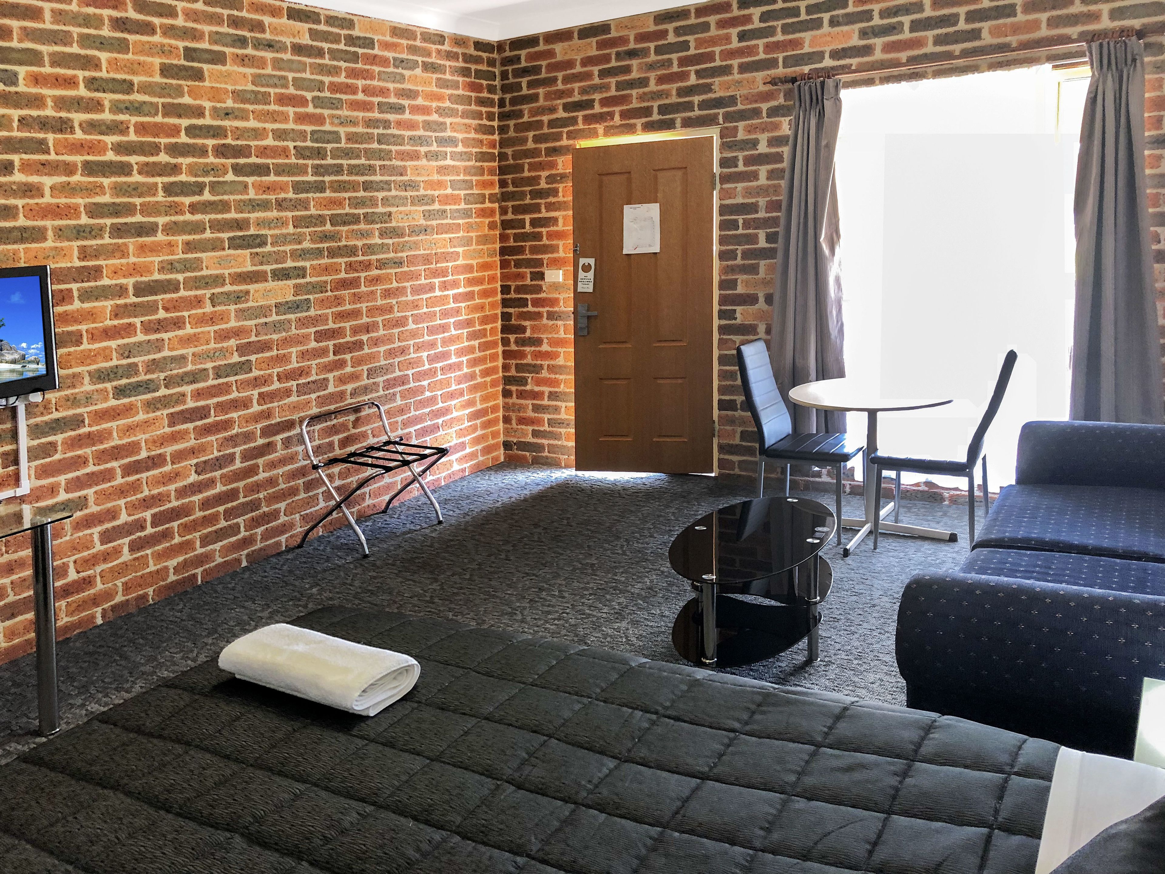 A hotel room with a brick wall, a bed with a white towel, a small table with chairs, and a TV on the wall.