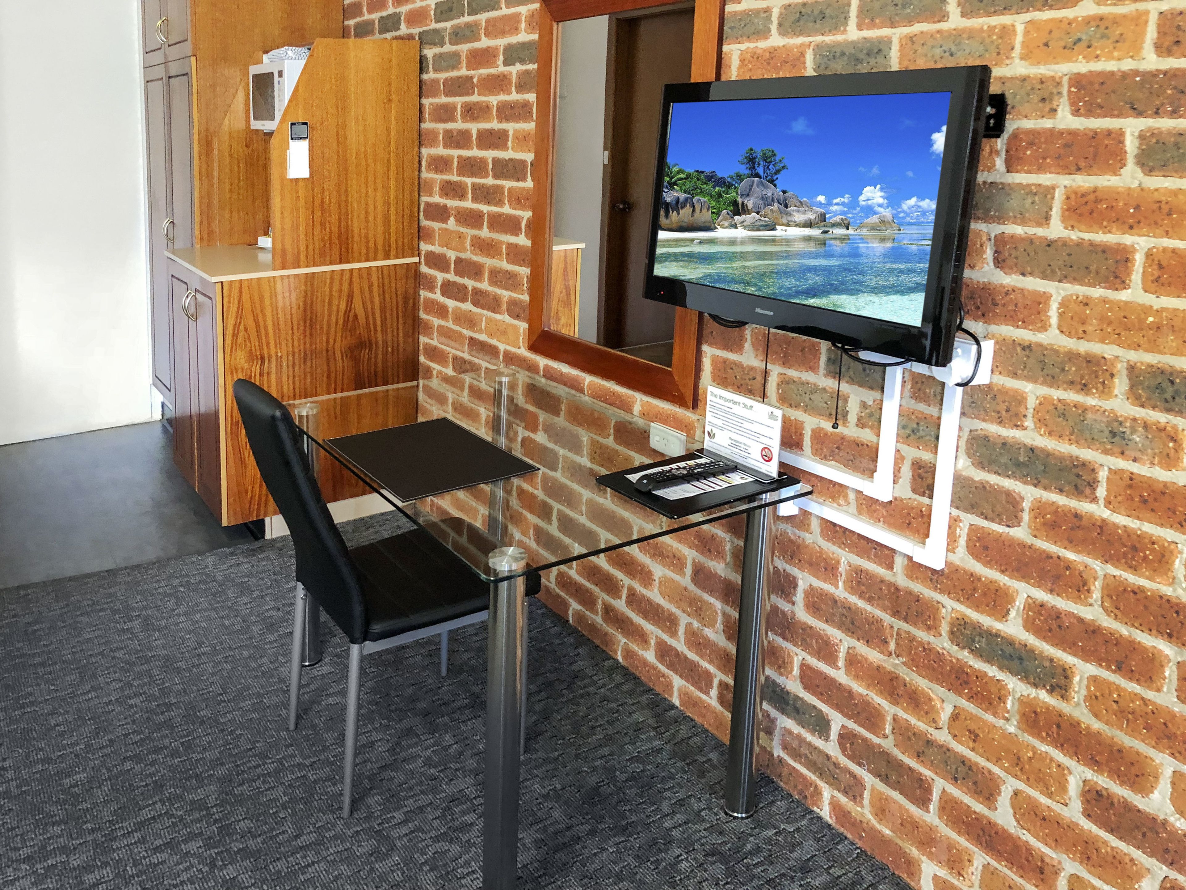 A room with a brick wall, a television mounted on the wall, a glass table, a black chair, and a wooden cabinet.