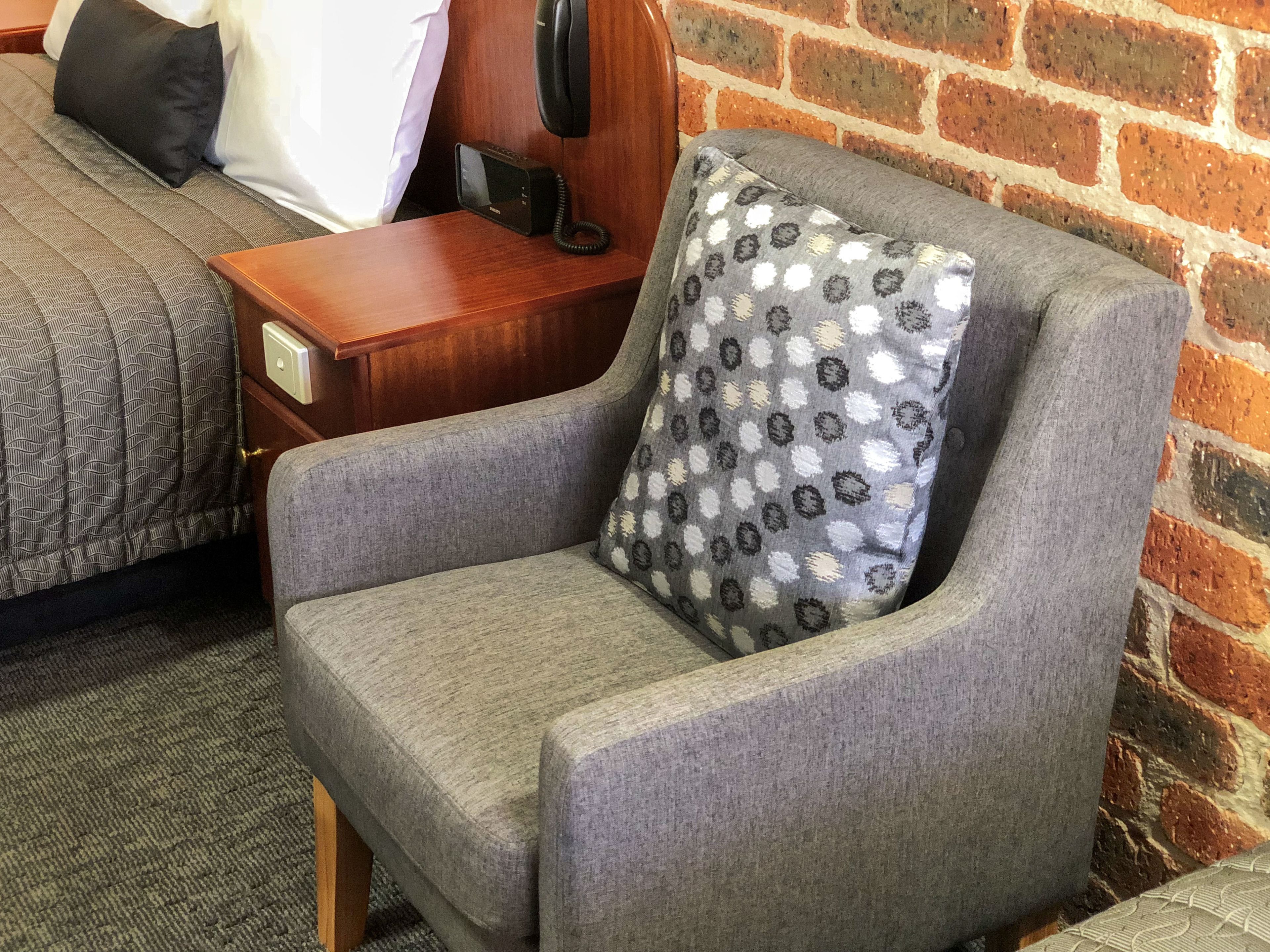 A grey armchair with a patterned pillow, a wooden nightstand with a black telephone, and a brick wall in the background.