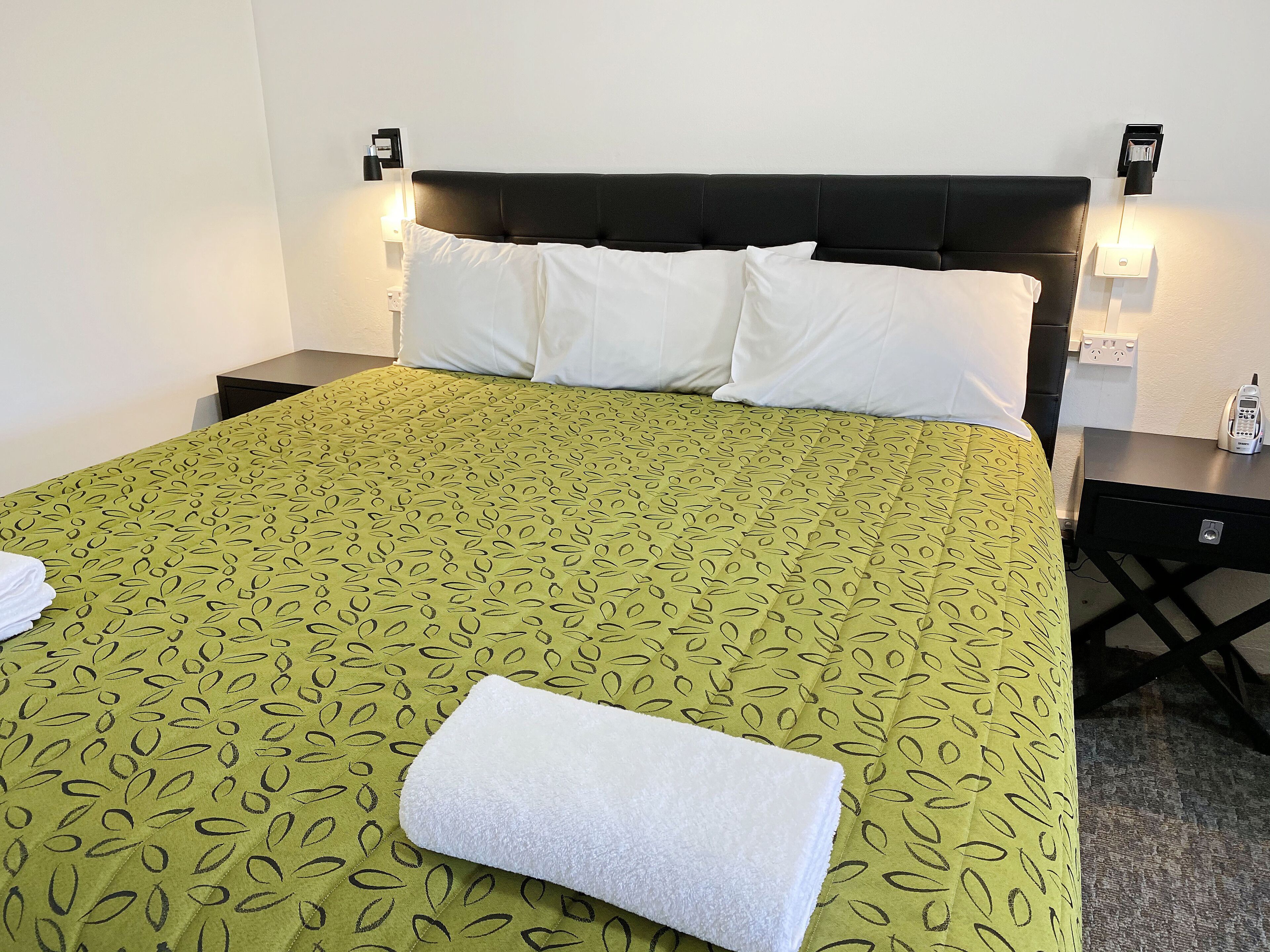 Una cama bien hecha con una colcha verde estampada, almohadas blancas y una toalla blanca doblada. Hay dos mesitas de noche, cada una con un teléfono y una lámpara.