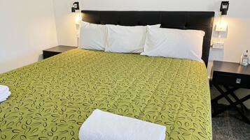 Una cama bien hecha con una colcha verde estampada, almohadas blancas y una toalla blanca doblada. Hay dos mesitas de noche, cada una con un teléfono y una lámpara.