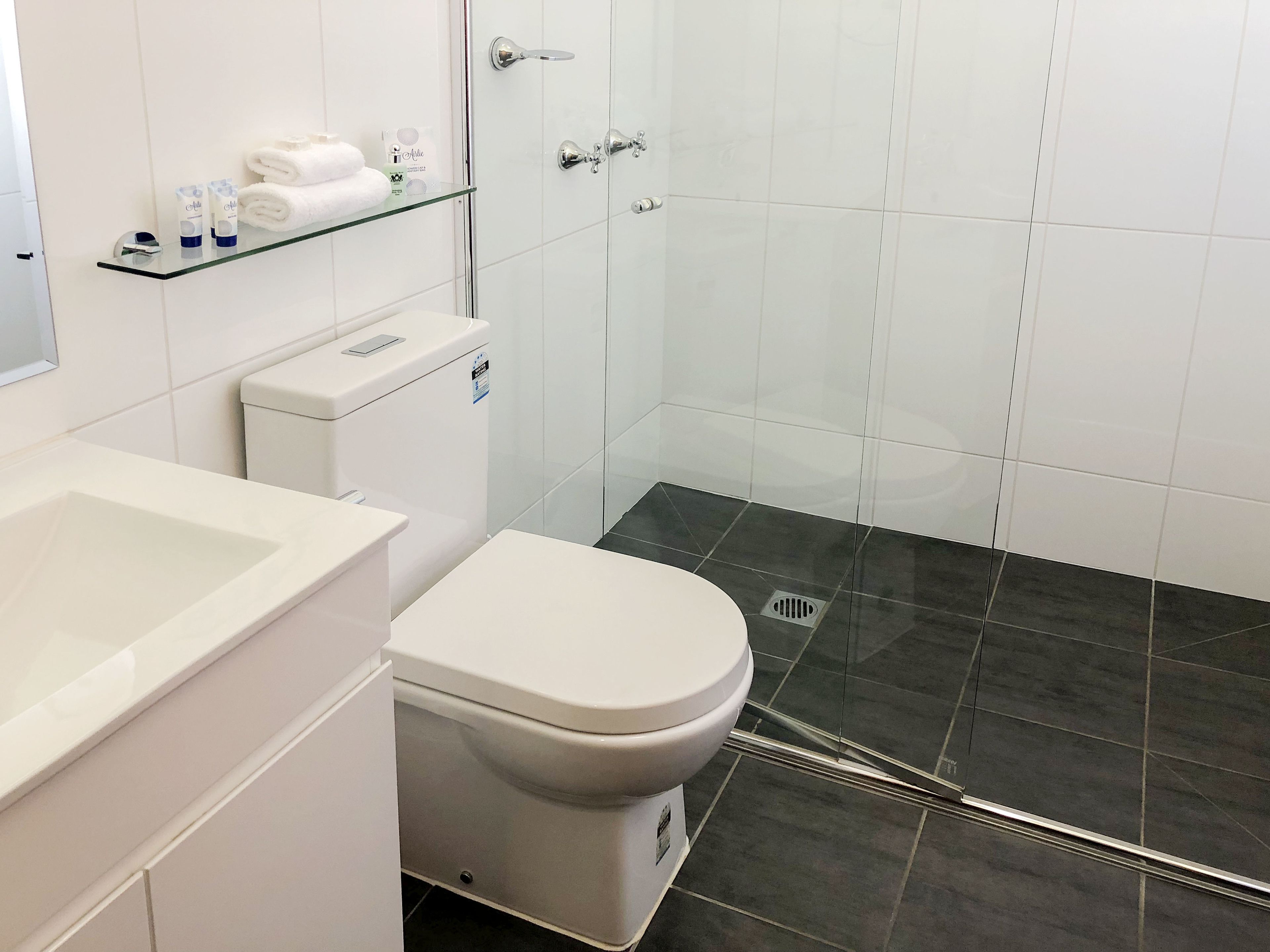 Et moderne badeværelse med et hvidt toilet, en hylde i glas med toiletartikler og en bruseniche.
