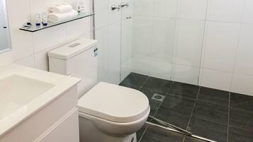 Et moderne badeværelse med et hvidt toilet, en hylde i glas med toiletartikler og en bruseniche.