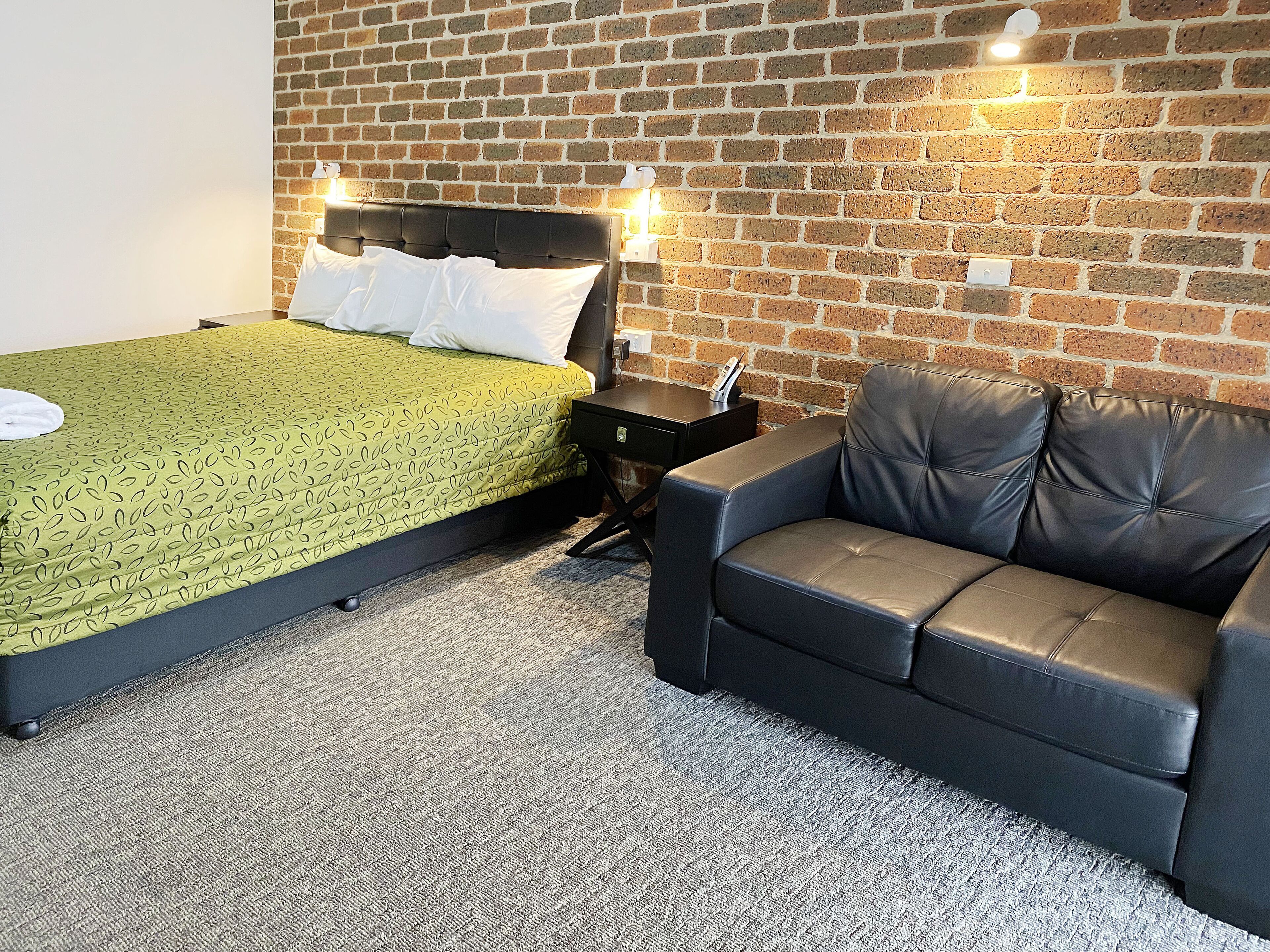 A hotel room with a bed, a leather sofa, a brick wall, and a carpeted floor.