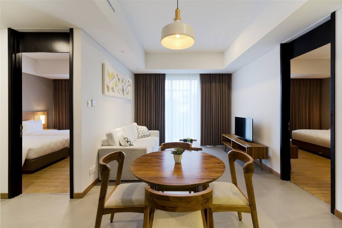 A modern hotel room with a bed, a sofa, a TV, a dining table with chairs, and a ceiling light.