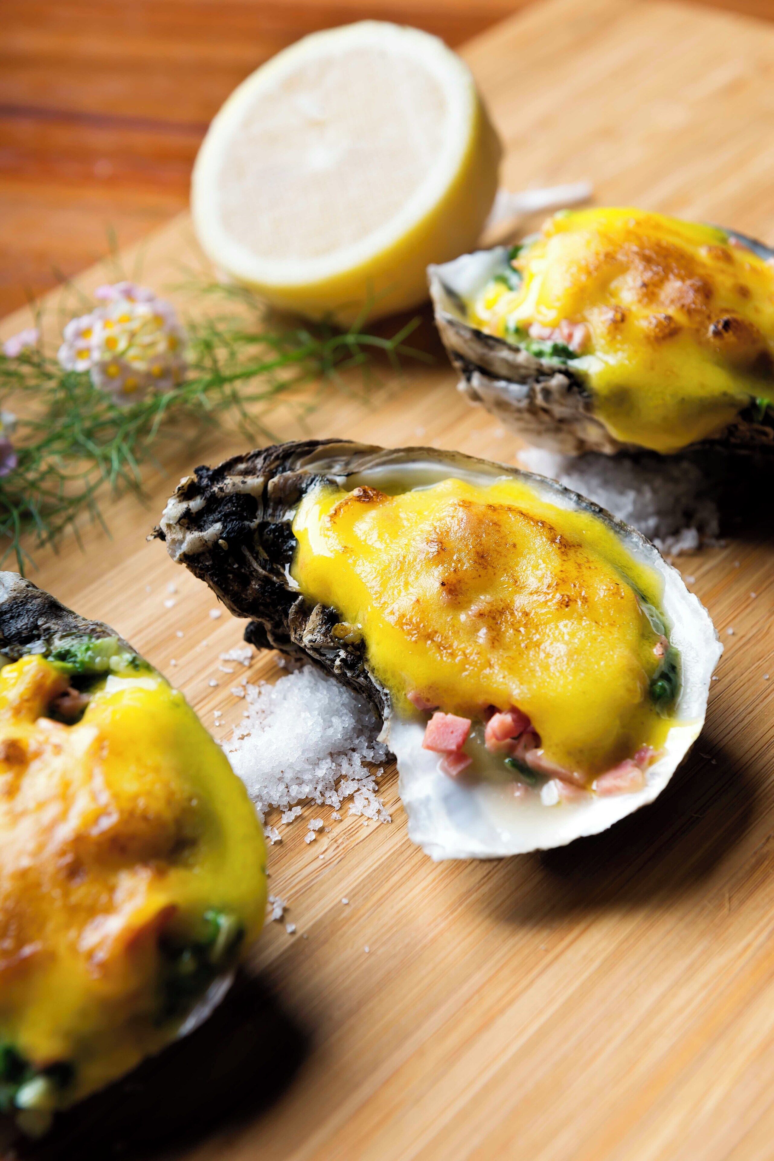A wooden cutting board with oysters topped with melted cheese and garnished with herbs.