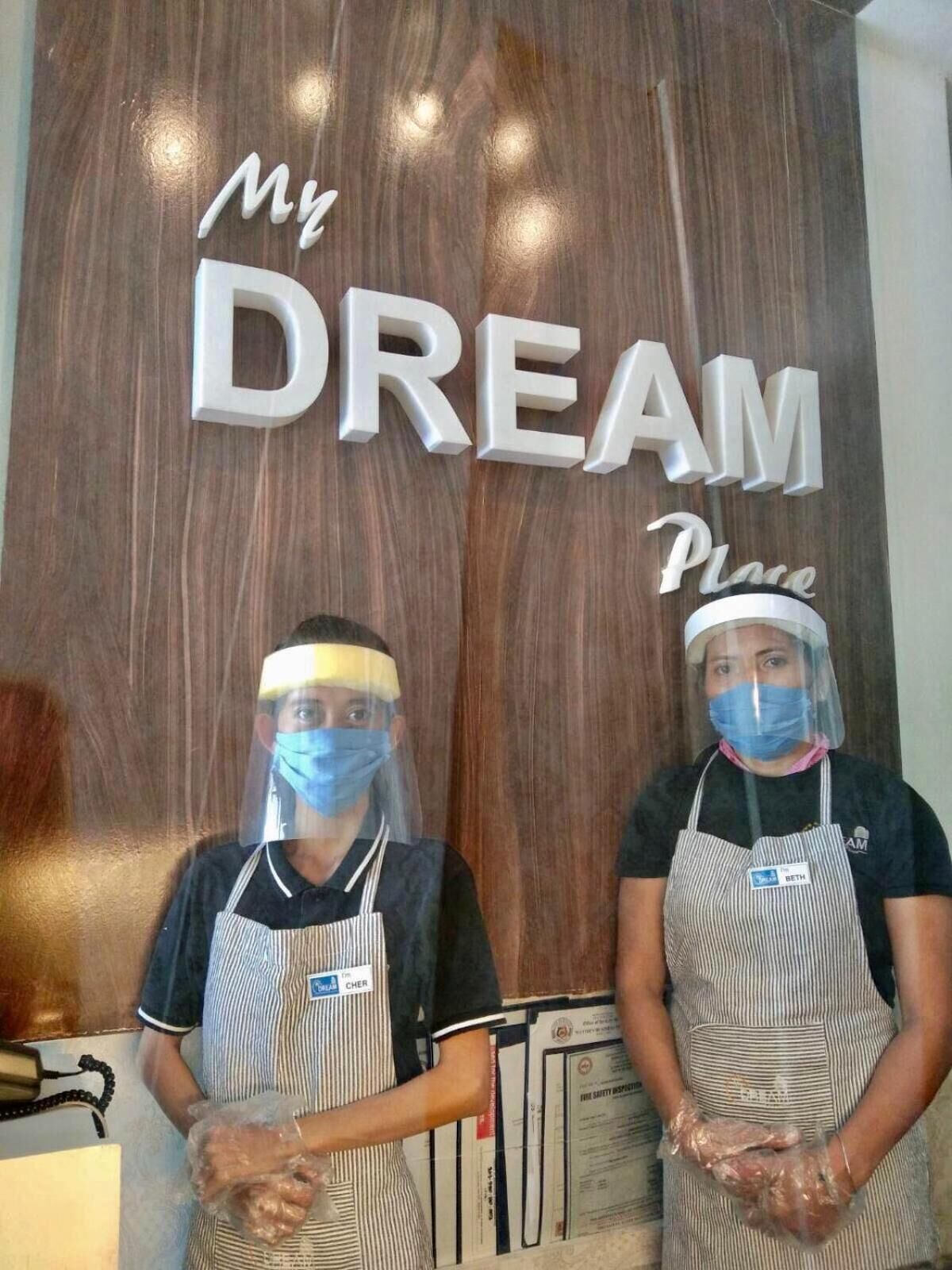 Dues persones portant delantals i protectors facials de peu davant d'un rètol de fusta que diu "My Dream Place".