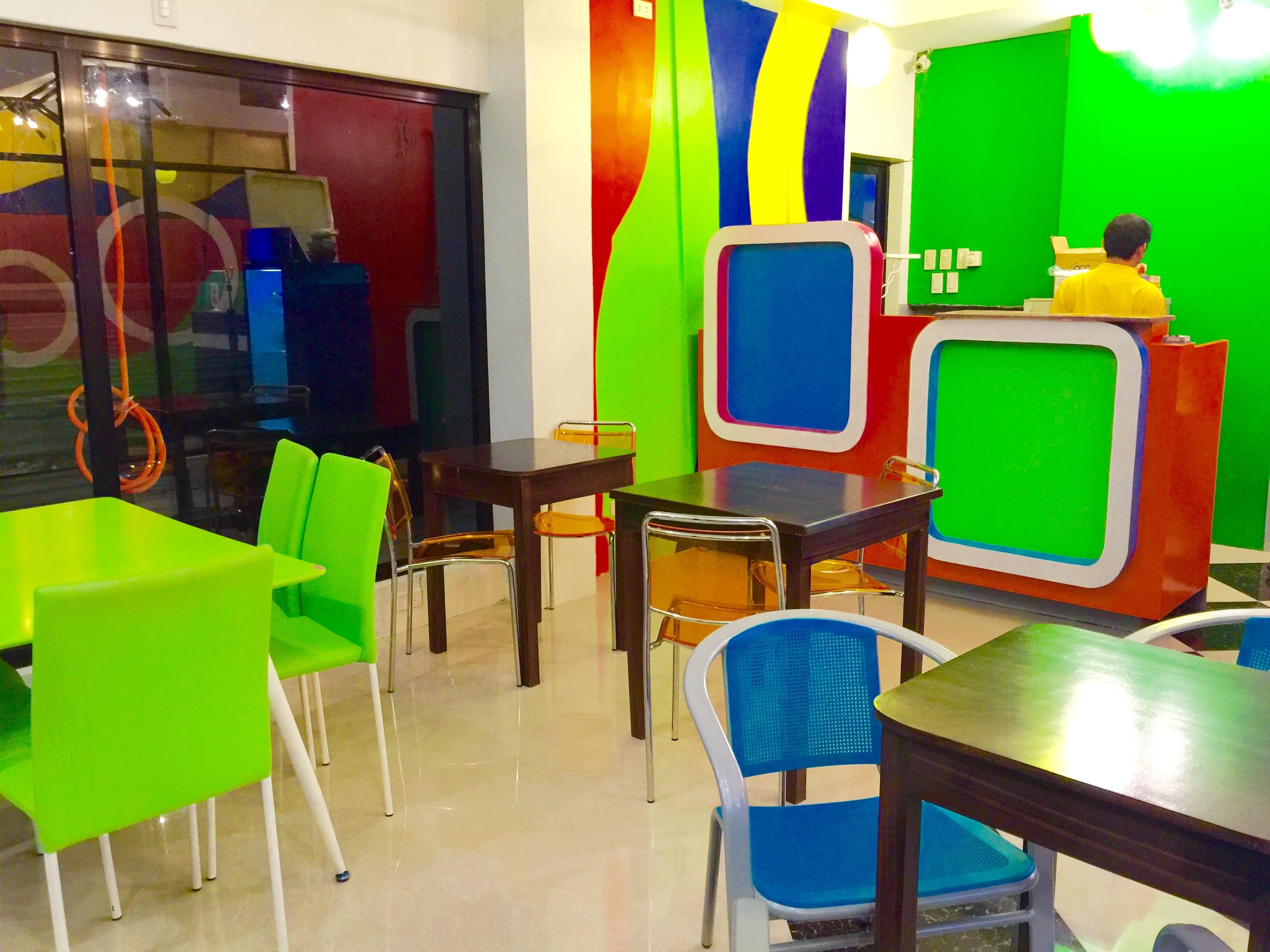 A brightly colored restaurant interior with green and blue chairs, a wooden table, and a green screen backdrop.