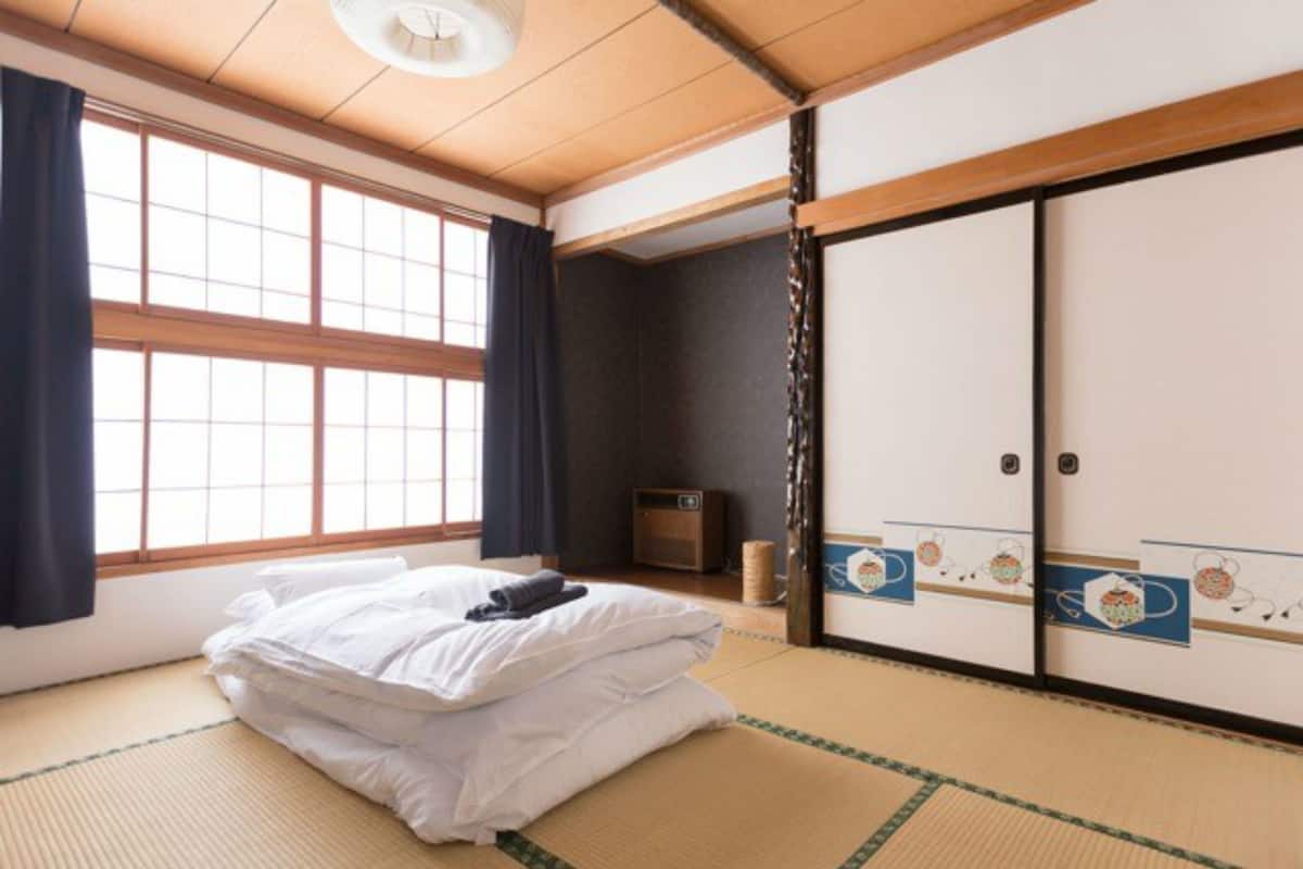 A traditional Japanese room with tatami flooring, a bed with white bedding, a futon, a sliding door with decorative patterns, and a small cabinet.