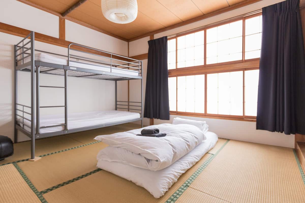 A room with tatami mat flooring, a bunk bed, a single bed with pillows, and a window with curtains.