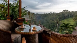 Un rincón de descanso de mimbre con una mesa de vidrio, dos sillones y vistas a un paisaje verde y exuberante.