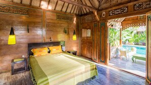 A wooden room with a bed, a small table, and a chair. There is a window with a view of greenery and a pool. - Hati Padi Cottages (Payangan)