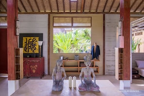 A room with a wooden ceiling, a large window with a view of greenery, a shelf with decorative items, and two statues on the floor. - Hati Padi Cottages (Payangan)