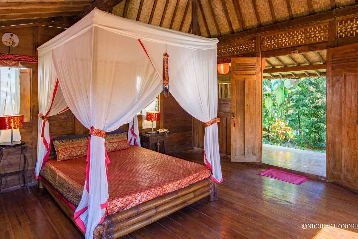 A four-poster canopy bed with a red patterned bedspread, wooden flooring, and a view of lush greenery outside.