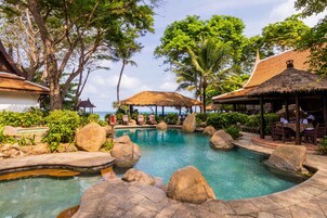 A pool surrounded by rocks and trees, with a thatched-roof structure and a dining area.