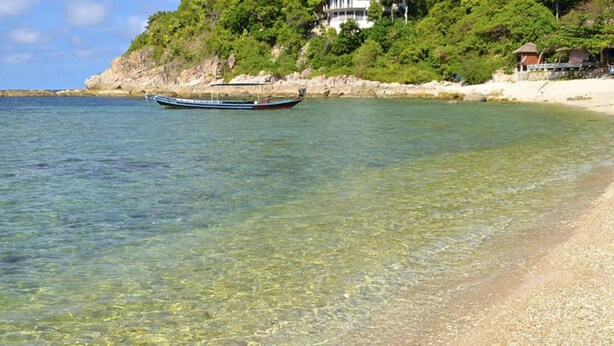 A clear beach with a boat in the water and a building on a hill in the background. - Bro&Sis Place (Koh Tao)