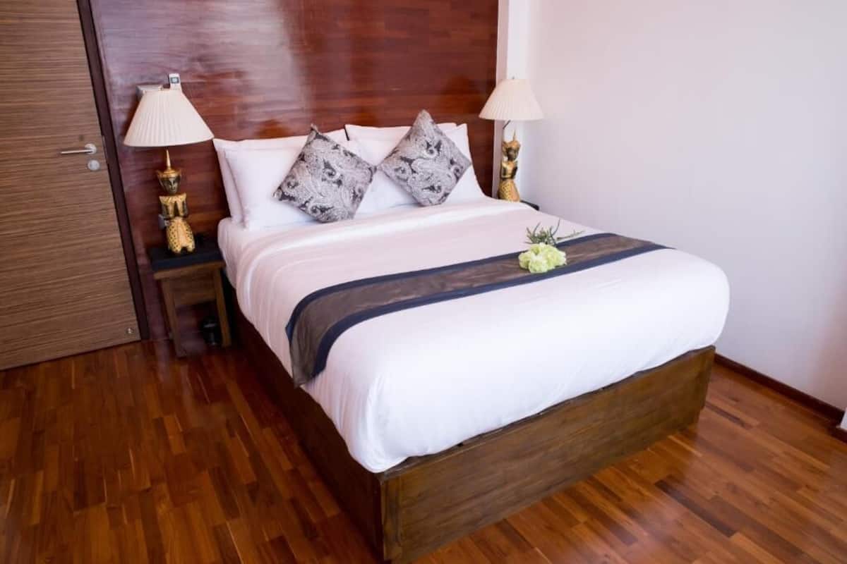 A neatly made bed with white linens and two decorative pillows, flanked by two bedside tables with lamps, in a room with wooden flooring and a wooden headboard.