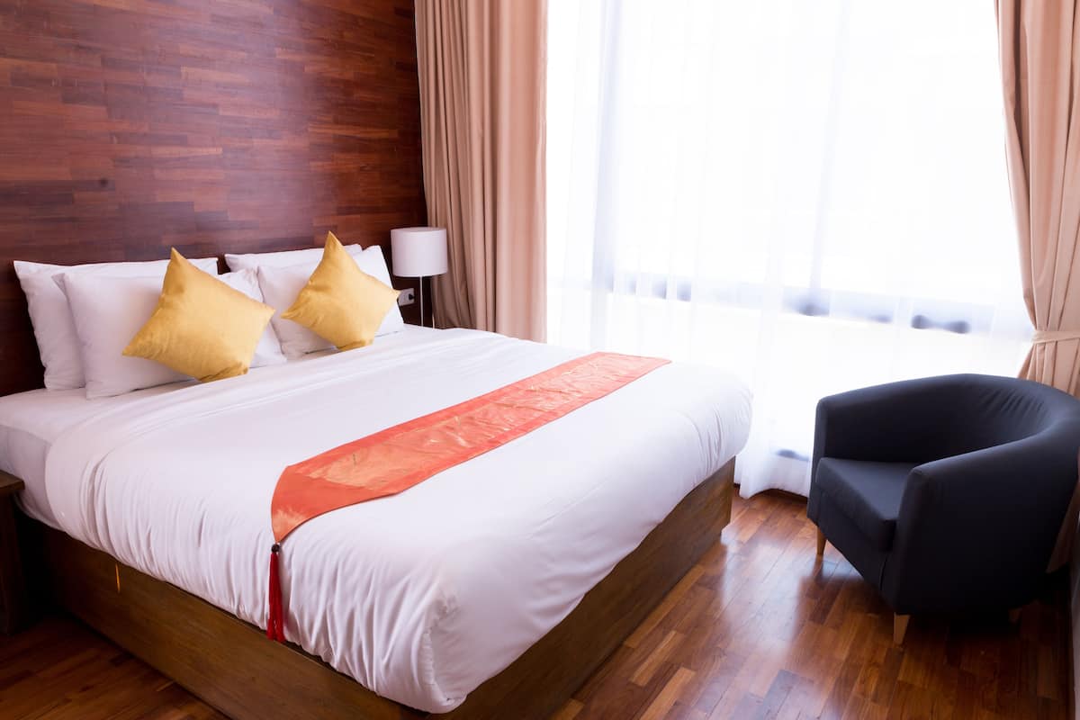 A hotel room with a wooden bed, white bedding, two yellow pillows, a red accent on the bed, a black armchair, a nightstand with a lamp, and a large window with curtains.