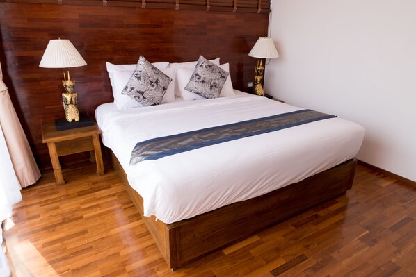 A neatly made bed with white bedding and patterned pillows, flanked by two bedside tables with lamps, in a room with wooden flooring and a wooden headboard.