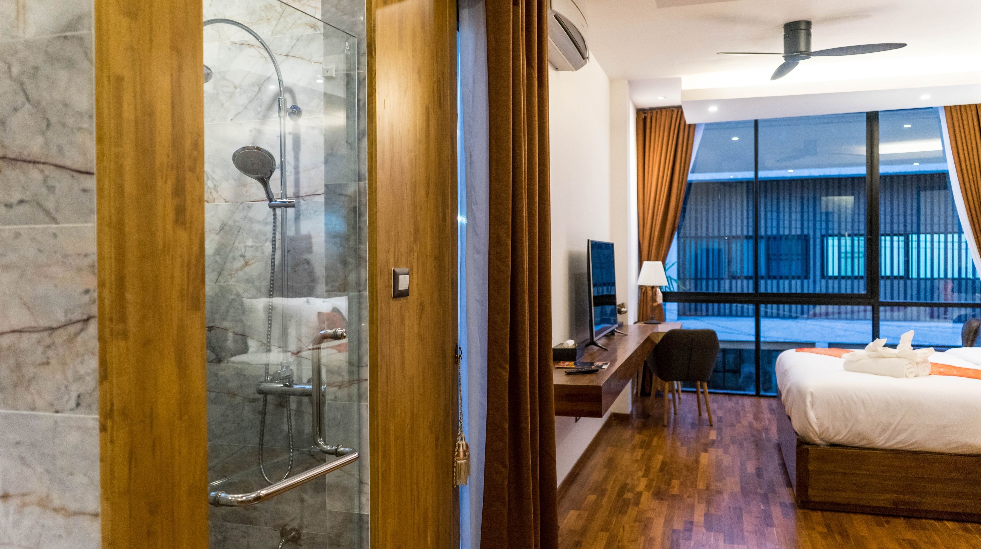 A hotel room with a glass-enclosed shower, a bed with a wooden headboard, a desk with a computer, and a large window with curtains.