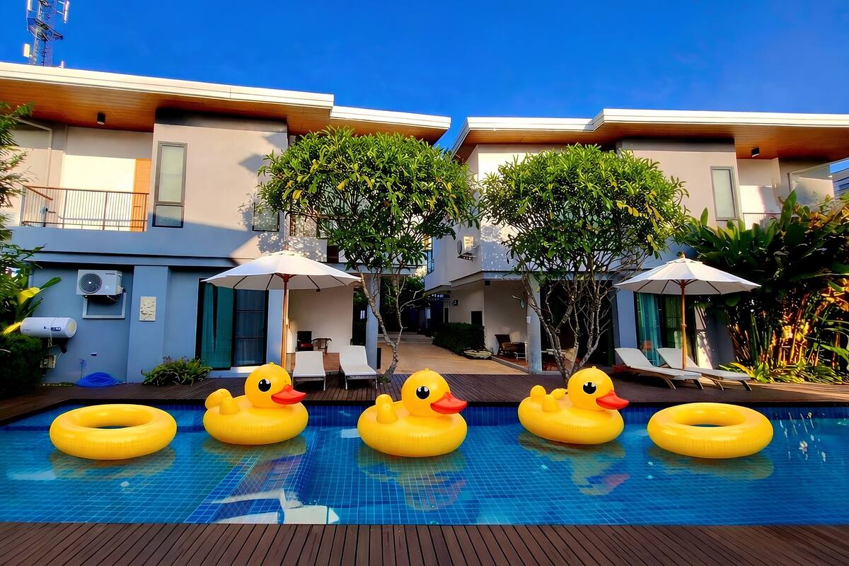 A swimming pool with inflatable duck floats, a covered seating area, and a building with balconies and trees.