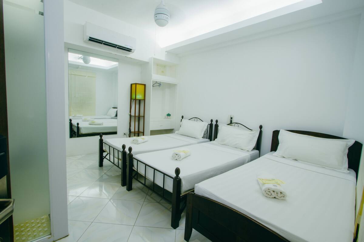 A hotel room with three single beds, a bathroom with a mirror, and a ceiling fan.