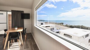 A room with a large window overlooking the sea, a wooden table with chairs, a flat-screen TV mounted on the wall, and a small plant on the table.