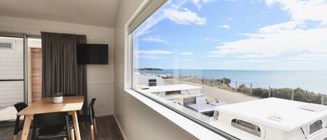 A room with a large window overlooking the sea, a wooden table with chairs, a flat-screen TV mounted on the wall, and a small plant on the table.
