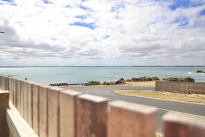 A coastal road with a wooden fence in the foreground, leading to a beach with calm waters and a clear sky.