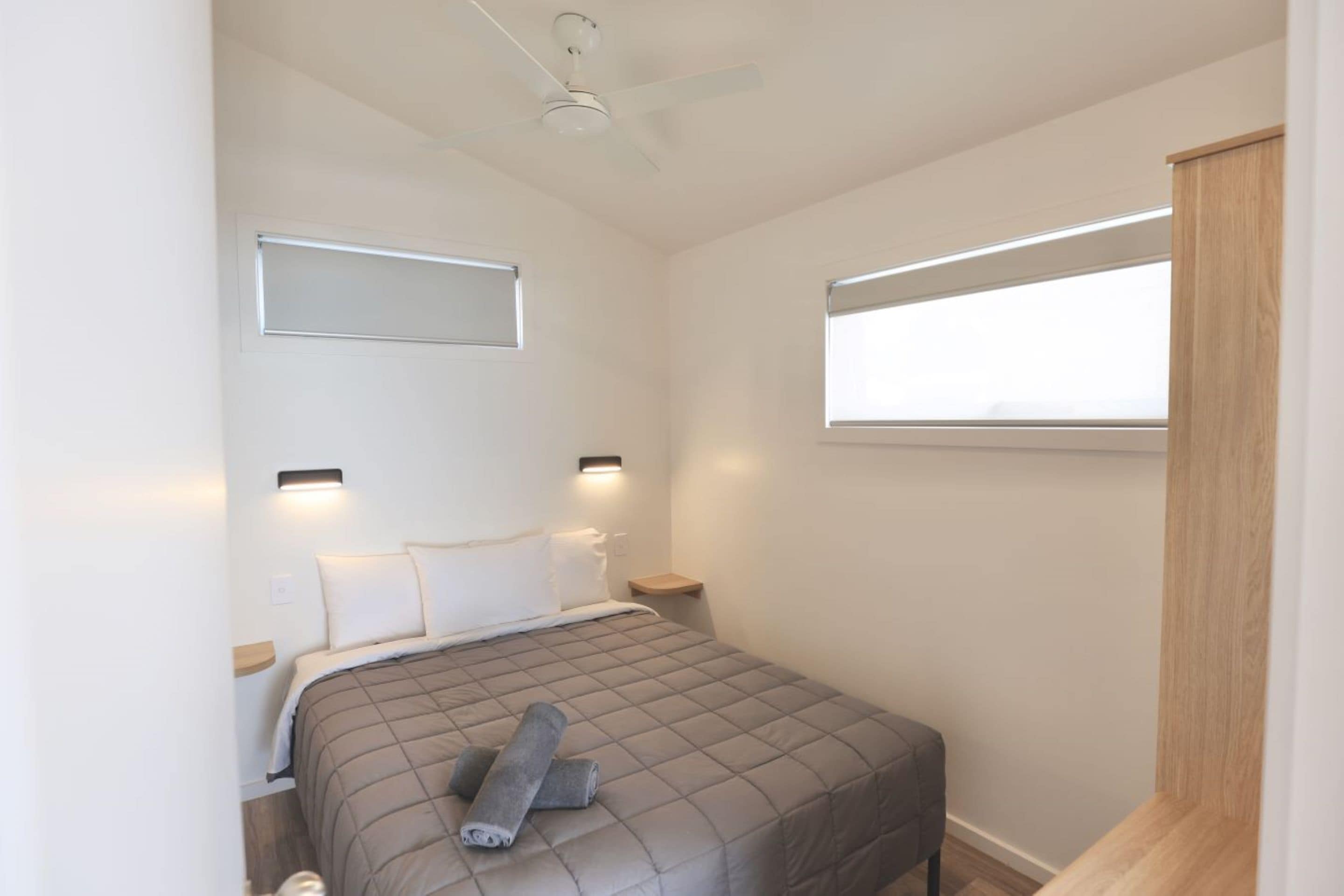 A bedroom with a bed, a ceiling fan, bedside lamps, a nightstand, and a window with blinds.