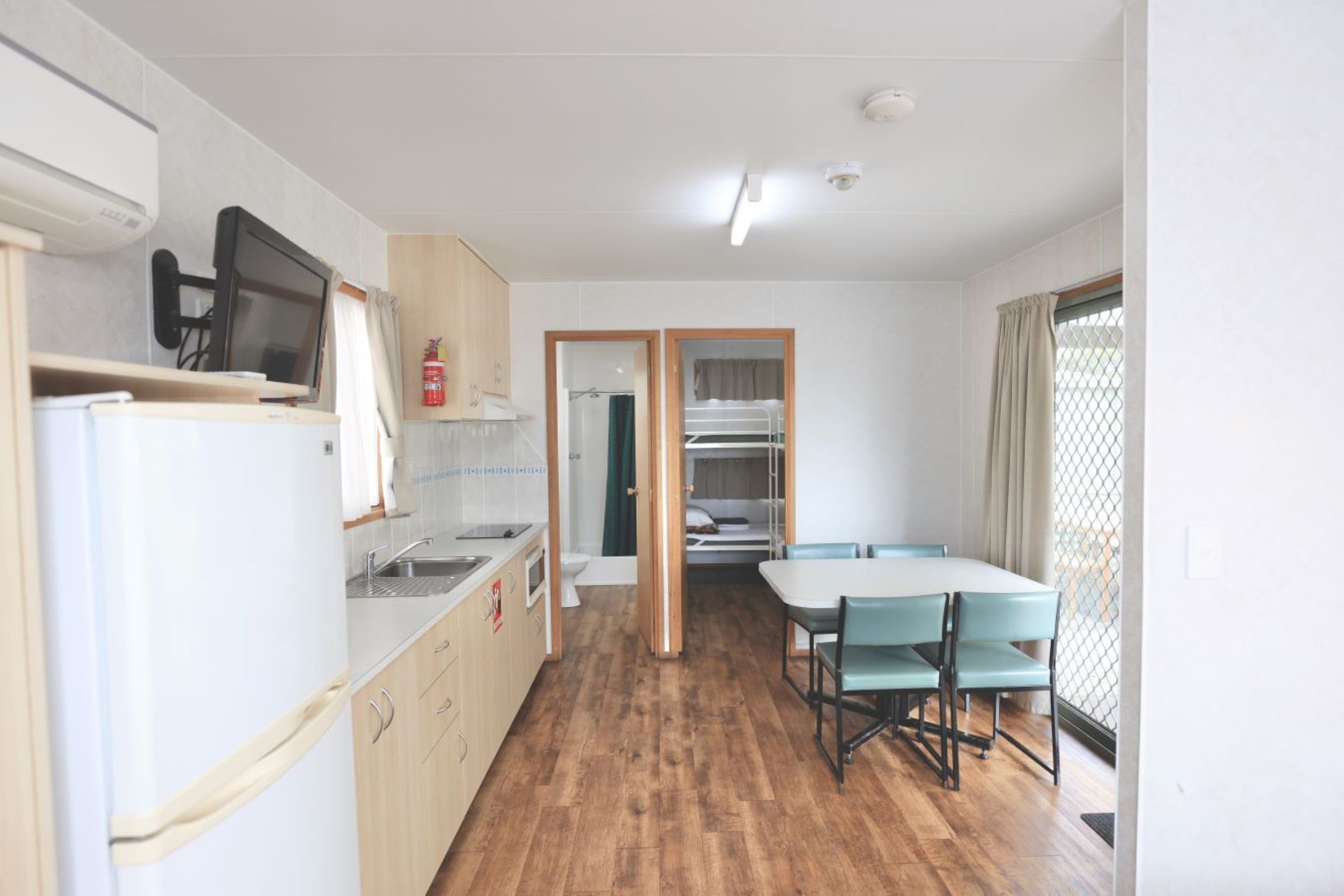 A compact kitchen with a refrigerator, microwave, and sink. There is a dining area with a table and chairs, and a view through an open door to a sleeping area with bunk beds.