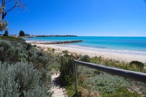 A coastal pathway leading to a sandy beach with clear blue waters, surrounded by vegetation and a wooden fence.