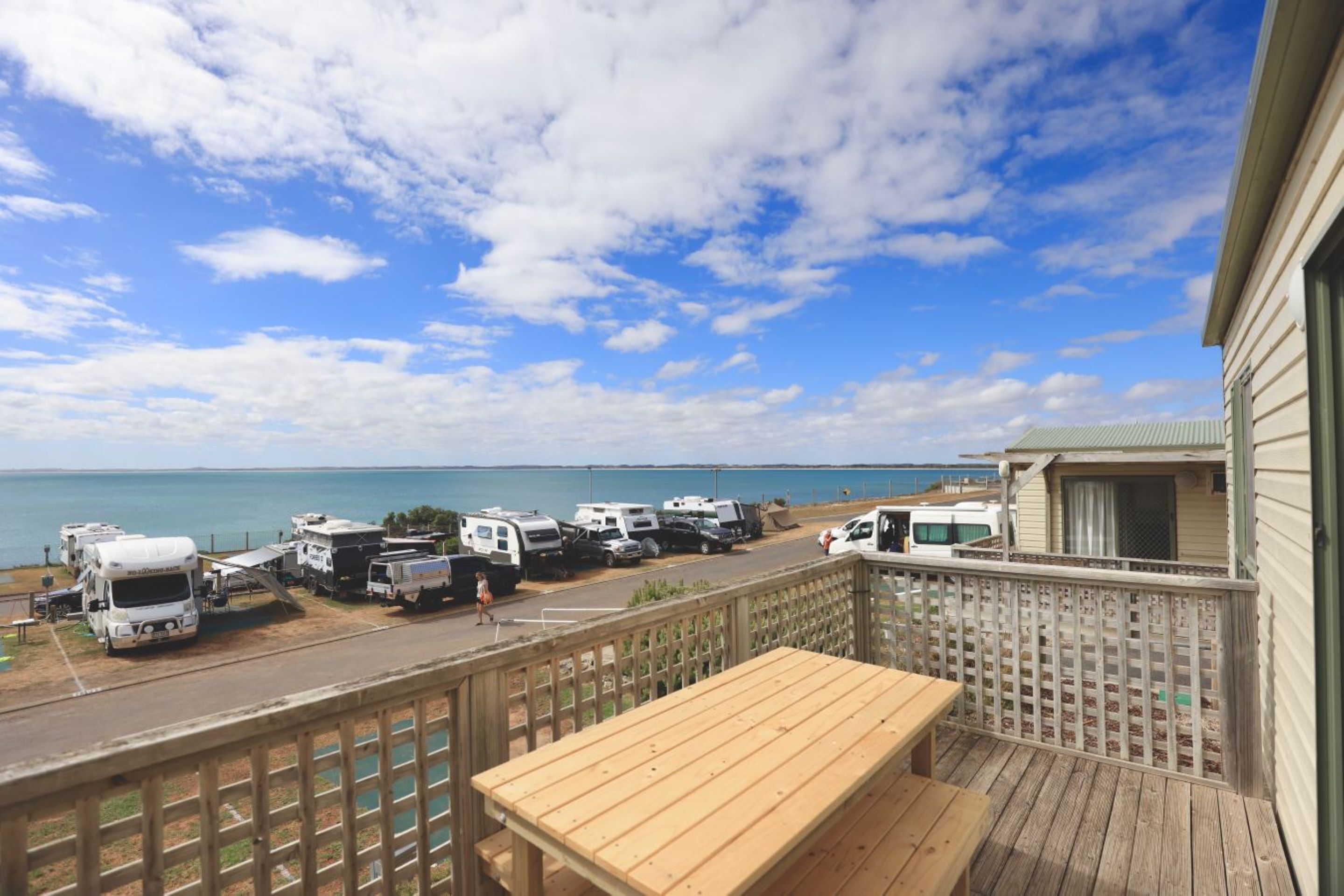 A deck with a wooden table overlooking a parking area with RVs and a view of the ocean.